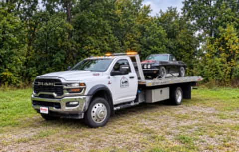 Safeway Services white Ram flatbed tow truck with a black classic Corvette loaded on the bed, parked on a tree-lined gravel road